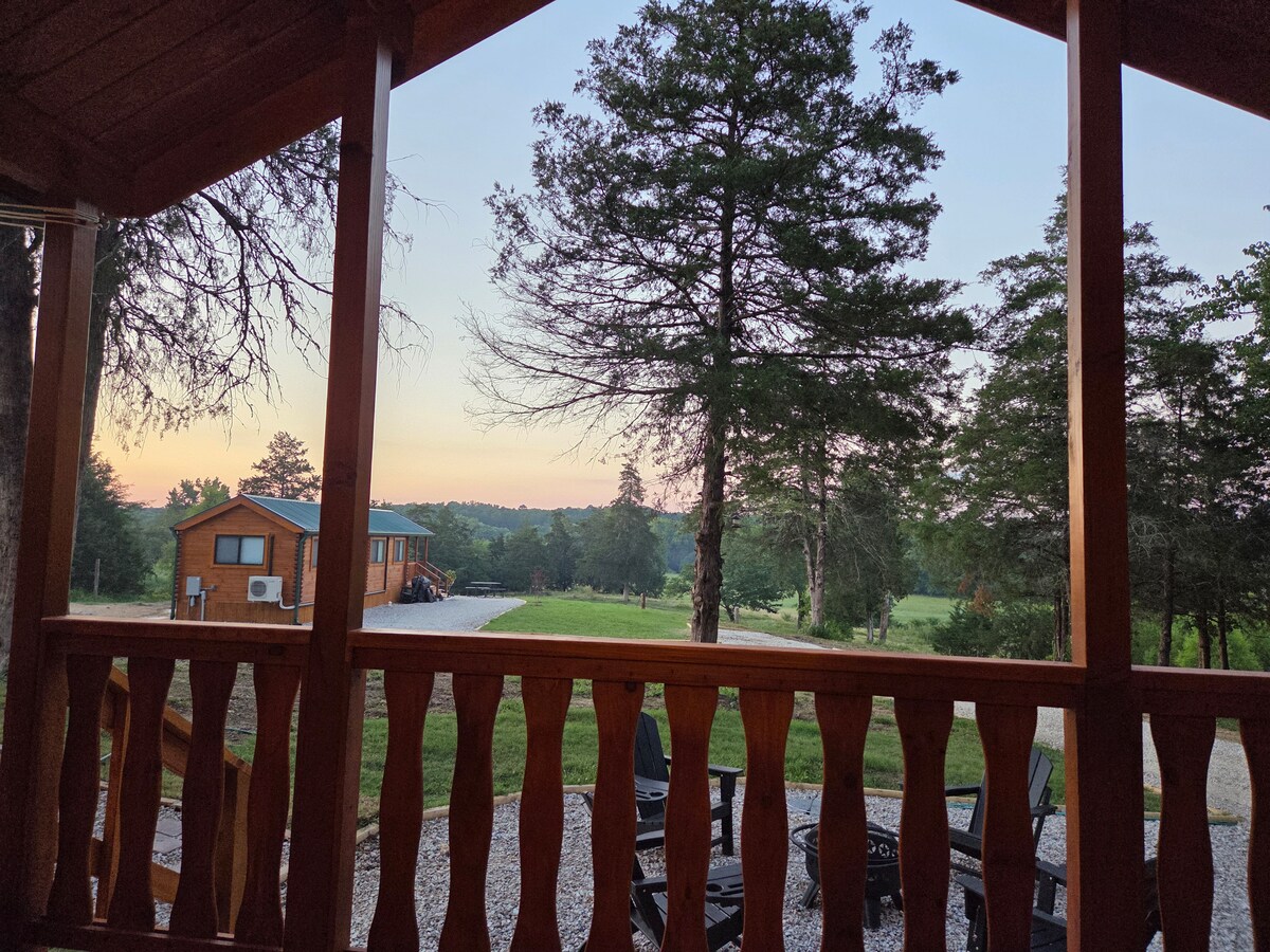 A view from the cabin's porch captures a serene landscape of rolling hills and trees under a soft morning sky. A neighboring cabin is visible in the distance, alongside a gravel pathway and outdoor seating arranged on the porch.