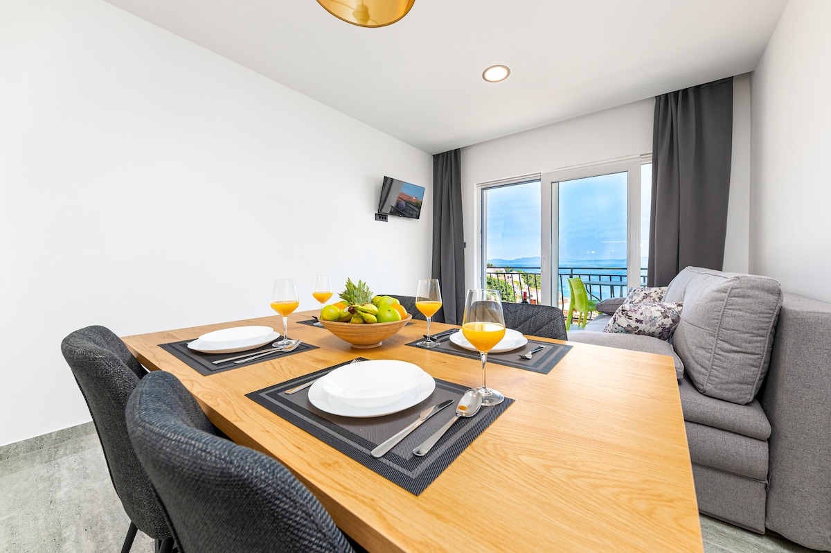 A dining area features a wooden table set for four, complete with glasses and a fruit bowl. A soft gray couch is positioned nearby, with large windows allowing natural light and offering views of the sea. A small television is mounted on the wall.