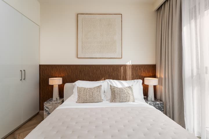 ห้องนอนที่มีสไตล์พร้อมเตียงนอนสบาย เครื่องเรือนทันสมัย และหน้าต่างสูงจากพื้นจรดเพดานที่มองเห็นวิวเมืองที่สวยงามทั้งกลางวันและกลางคืน
