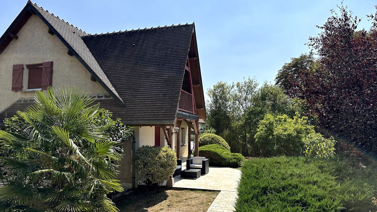 The exterior view of the villa showcases a well-maintained garden surrounded by lush greenery. The house features a sloped roof and attractive wooden details. Outdoor seating is visible on the patio, providing a relaxing space to enjoy the surrounding nature.