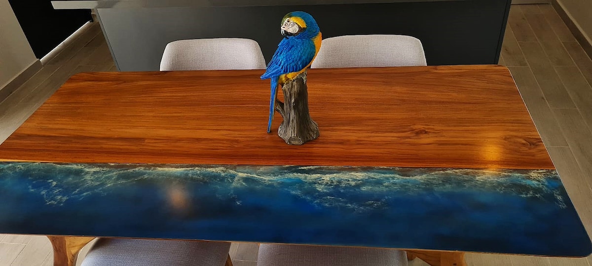 A wooden dining table is featured, showcasing a unique design with a blue ocean-themed section at the edge. In the center, a decorative parrot sculpture adds a vibrant touch, while two light-colored chairs accompany the table, creating a cohesive dining space.