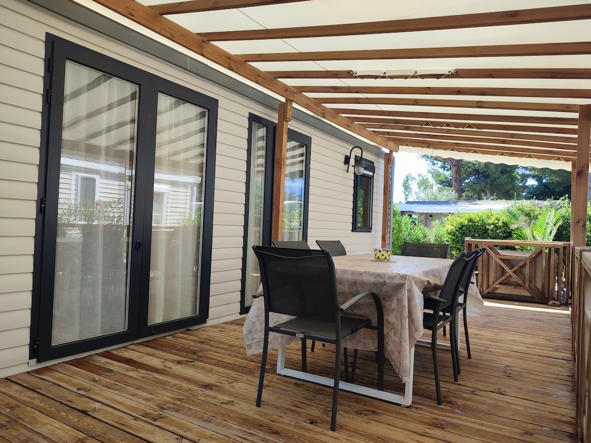 A shaded outdoor terrace features a wooden deck with a large table and several chairs arranged for dining. Large glass doors lead into the interior space, while greenery is visible in the background, providing a serene atmosphere.