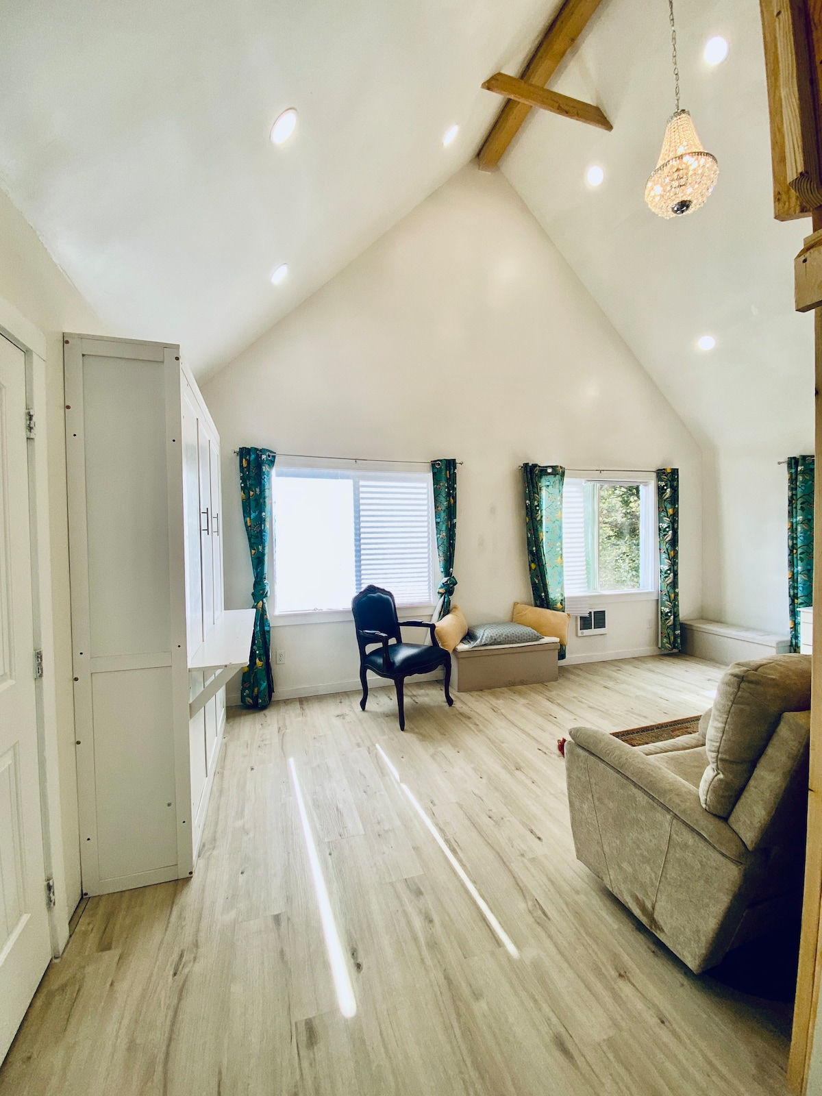 A bright room with a high ceiling is enhanced by natural light streaming through two windows, adorned with patterned curtains. A comfortable chair is placed beside a window, while a cozy seating area is located near the wall. Light-colored flooring complements the overall space.