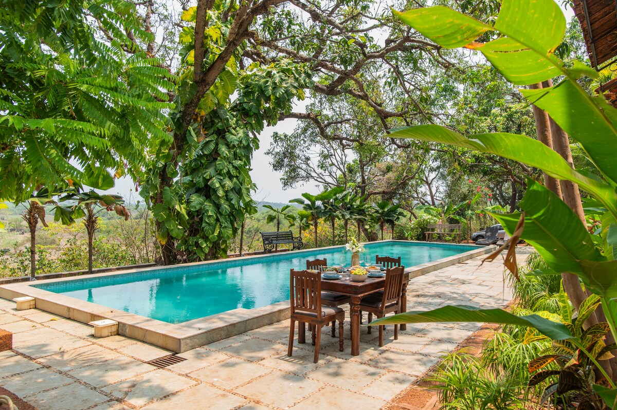 A serene pool area is framed by lush greenery, with two long pools reflecting the surrounding trees. Wooden dining furniture is positioned nearby, inviting outdoor meals amidst nature. The stone patio offers ample space for relaxation, enhancing the tranquil environment.