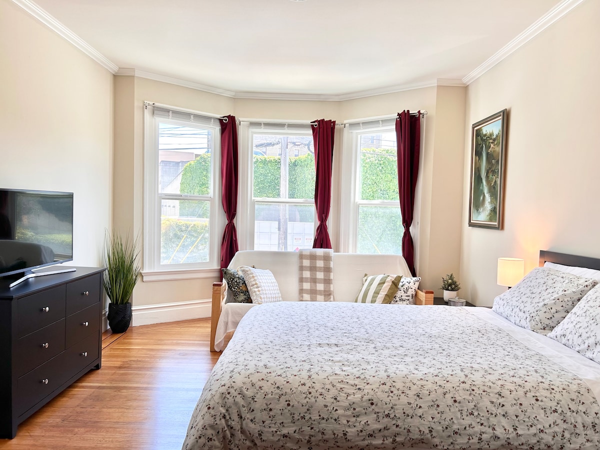 A spacious bedroom is presented with a comfortable bed dressed in floral bedding. A natural light-filled bay window, framed by burgundy curtains, overlooks greenery outside. A television is positioned on a dresser, and a cozy seating area is arranged with soft throws.