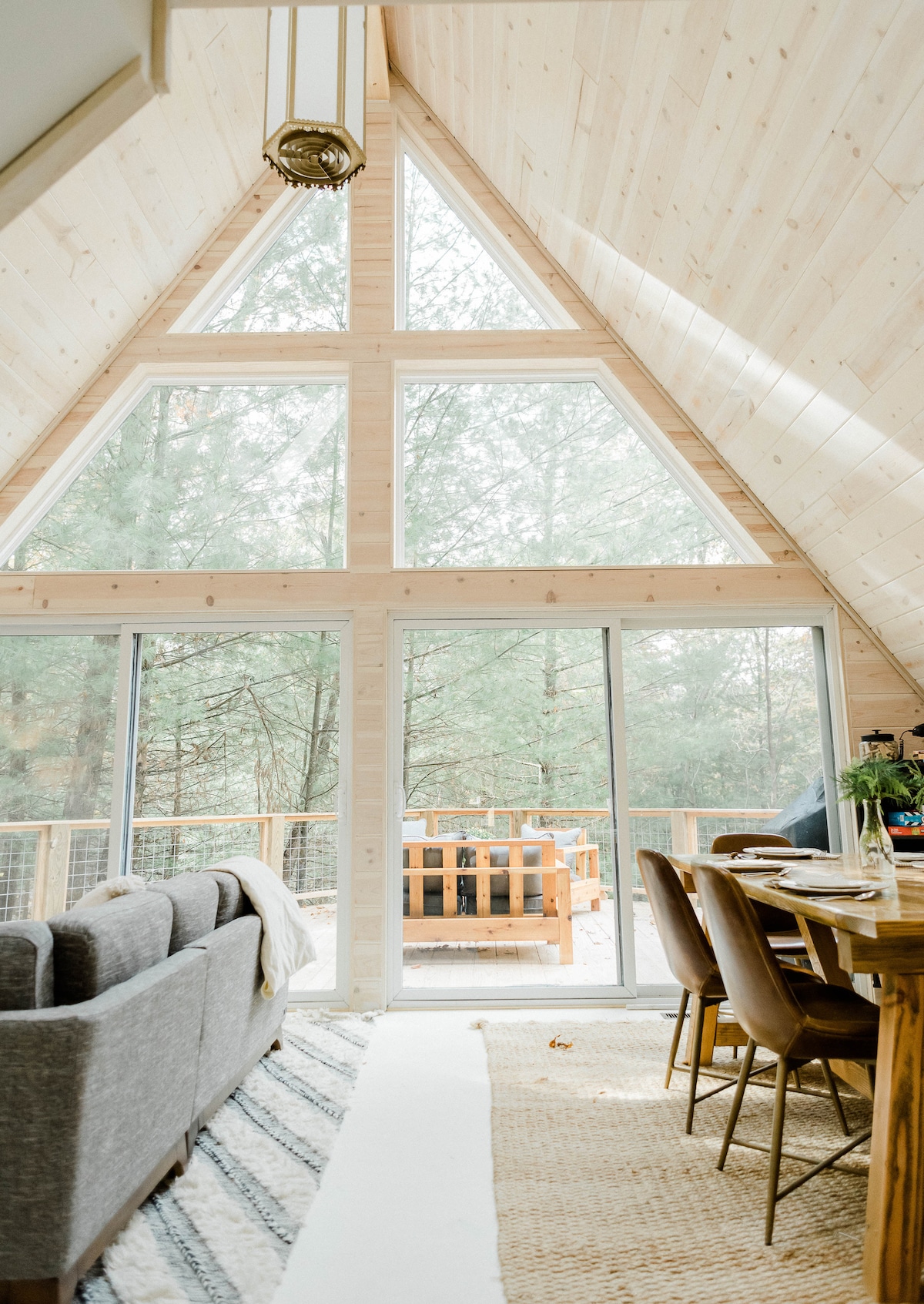 The airy living space features a high A-frame ceiling with large windows providing views of the surrounding trees. A sectional sofa and a wooden dining table are positioned to make the most of the natural light, creating an inviting space for gatherings.