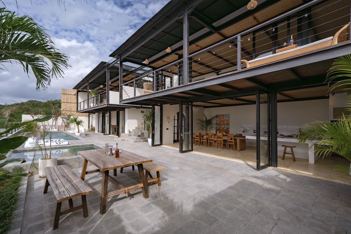 An expansive outdoor patio is visible, featuring a wooden dining table surrounded by benches. The area is framed by lush greenery and overlooks a spacious pool. Large glass doors provide a seamless connection between indoor and outdoor spaces, enhancing the villa's open feel.