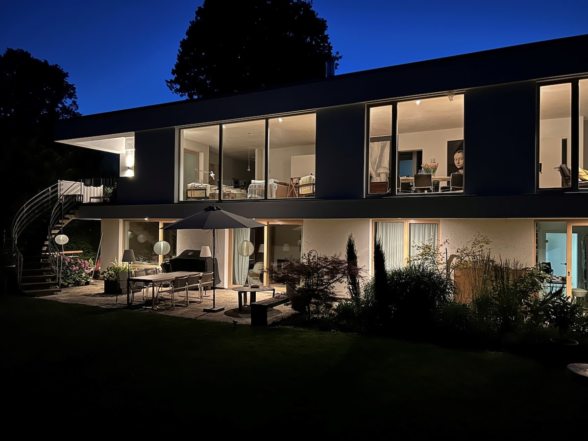 The exterior of the apartment is illuminated at night, showcasing large glass windows with soft interior lighting. A spiral staircase leads to the upper level, while a patio area is furnished with tables and chairs under an umbrella, surrounded by well-maintained gardens.