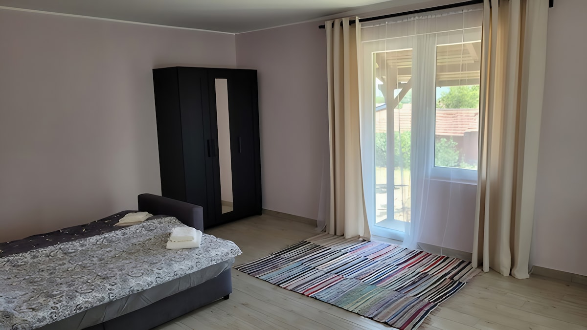 A spacious room features a cozy bed dressed in grey bedding, complemented by neatly folded towels. A modern wardrobe with mirrored doors stands against a light purple wall. Natural light filters through sheer curtains over a window, illuminating a colorful striped rug on the wooden floor.