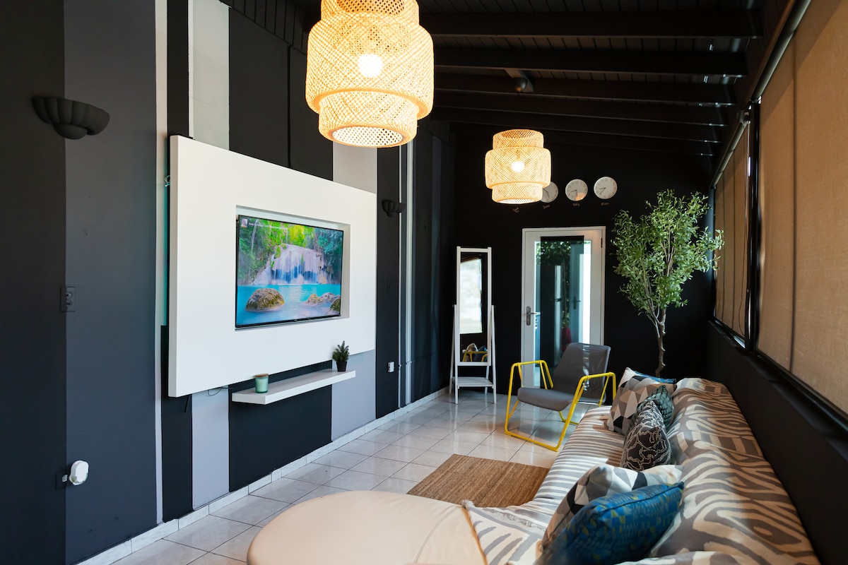 A stylish living area is designed with neutral tones complemented by a striped sofa. Modern hanging light fixtures create a warm glow, while a large television is mounted on a white wall. A potted plant and colorful chair add a touch of vibrancy to the space.