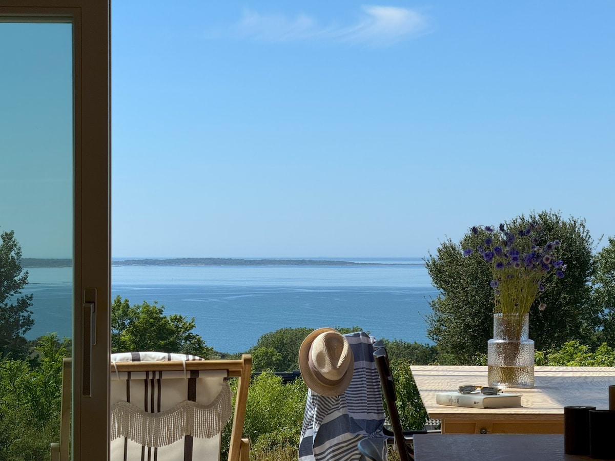 A bright view from the open doors showcases the calm blue sea and distant horizon. The outdoor dining space features a wooden table set with a vase of flowers and a sunhat resting atop a striped blanket. Nearby, two comfortable chairs are positioned to enjoy the scenery.