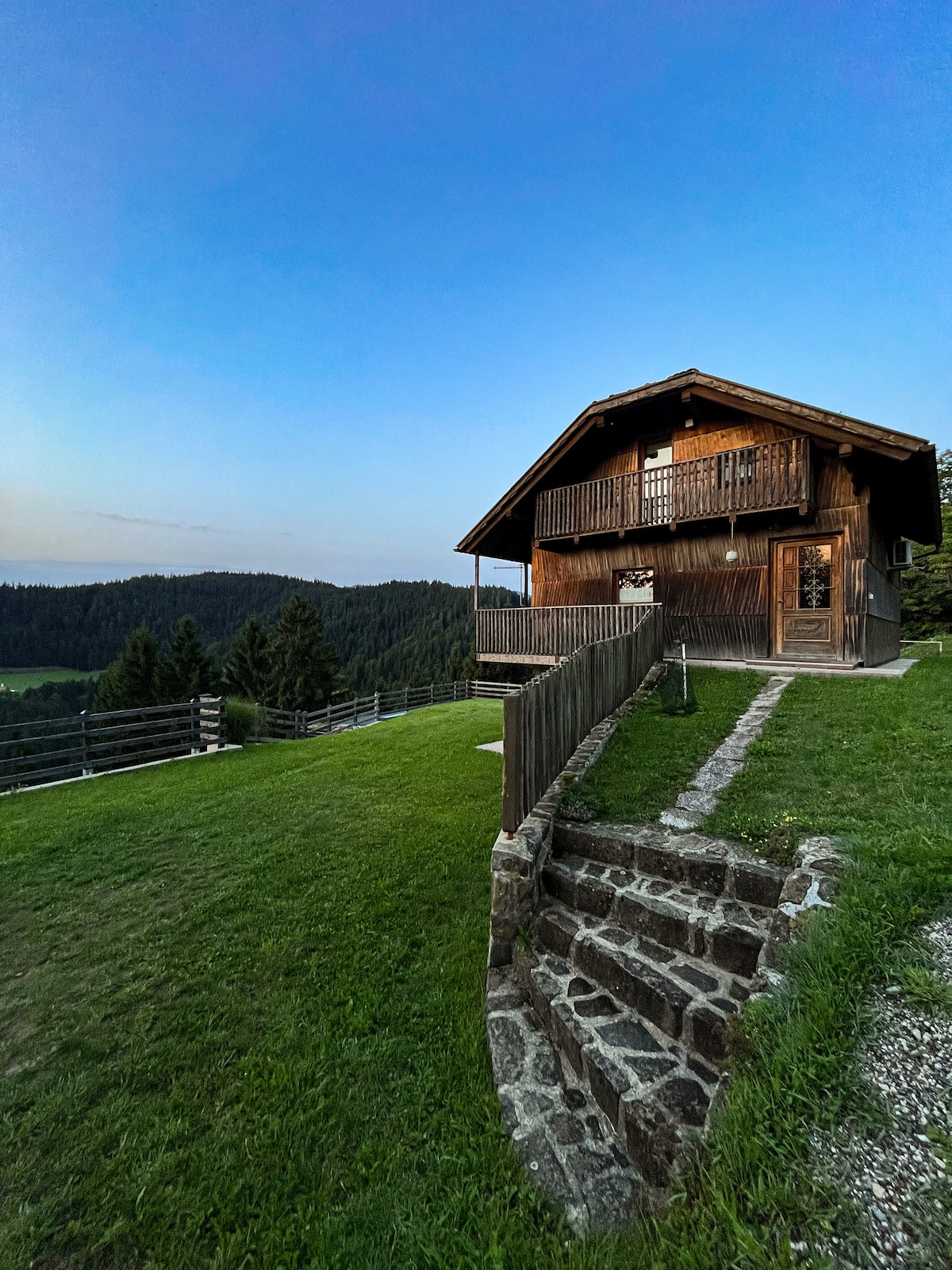 A charming wooden house is nestled in a green landscape, with a balcony overlooking the serene surroundings. Stone steps lead from the yard to the entrance, framed by lush grass and natural foliage, set against a clear blue sky.