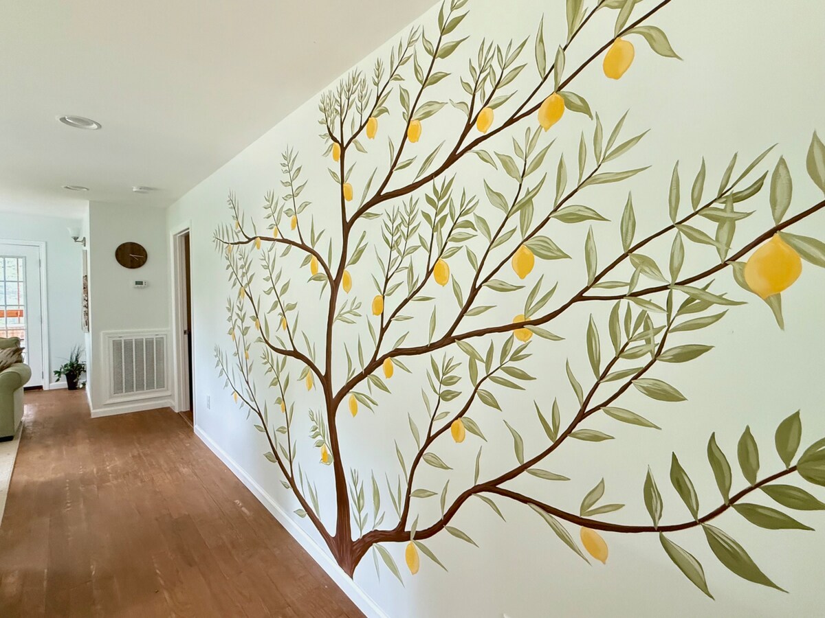 A vibrant mural depicting a lemon tree adorns the wall, featuring detailed green leaves and yellow lemons. The surrounding wall is a soft neutral color, complementing the warm wood flooring, which stretches down a hallway leading to other areas of the home.