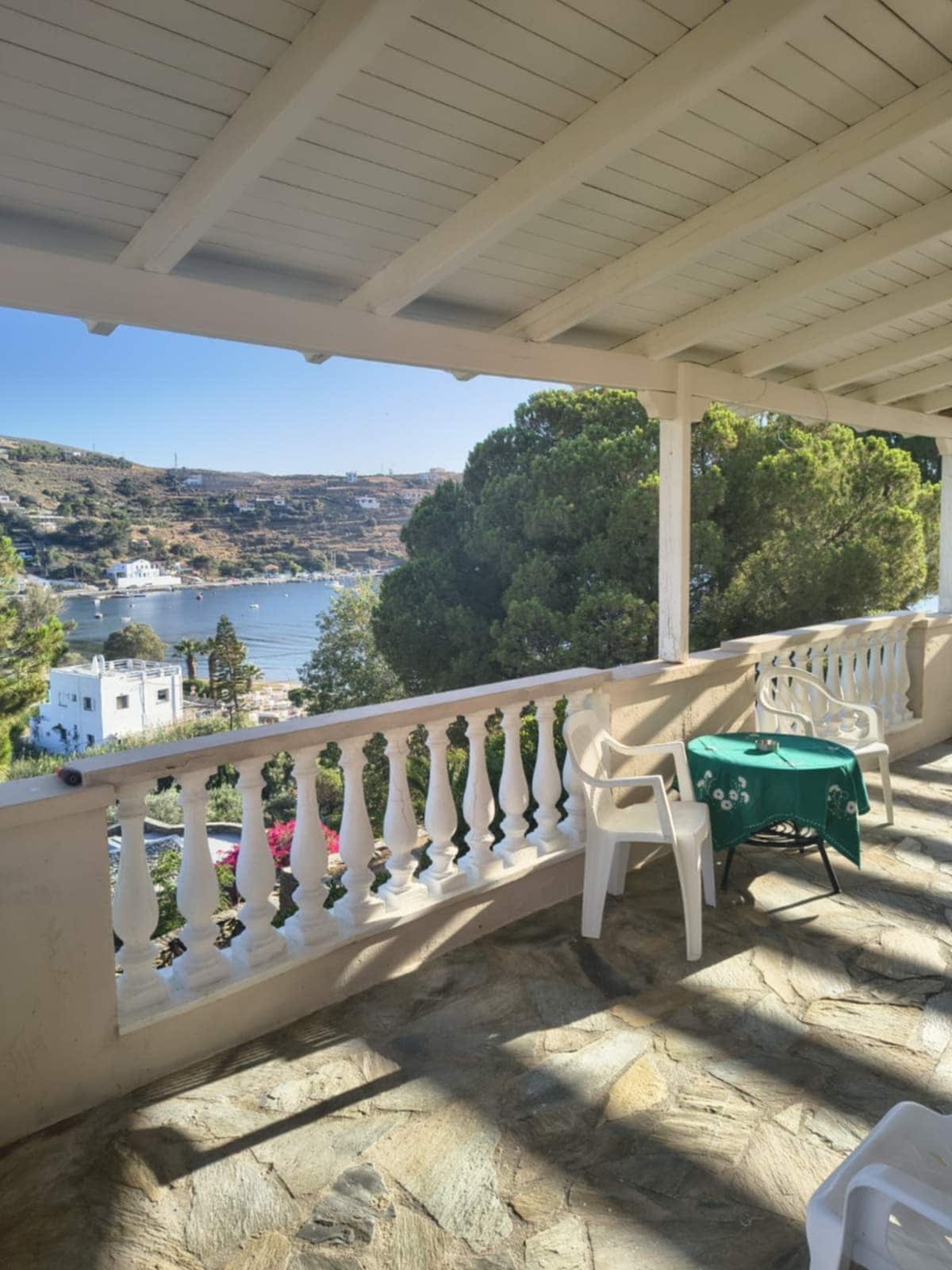 A balcony features a stone floor and white railings, overlooking serene water views and greenery. A small green table is accompanied by two white chairs, providing a cozy outdoor seating area. Sunlight casts gentle shadows, creating a welcoming atmosphere.