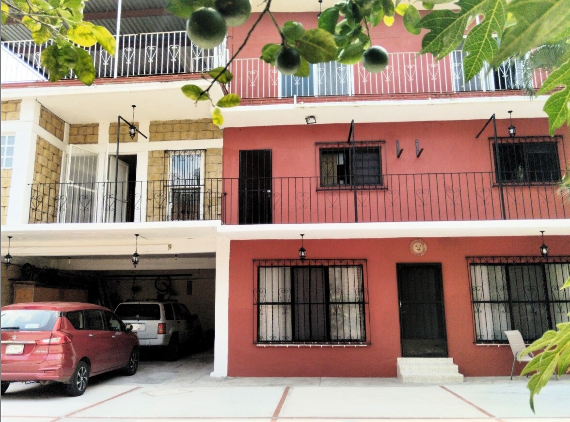 The building features a two-level design with a combination of stone and brick facades. A covered parking area accommodates vehicles, showcasing convenient access. Balconies with metal railings are visible on the top level, while the ground floor displays large windows covered by light curtains.