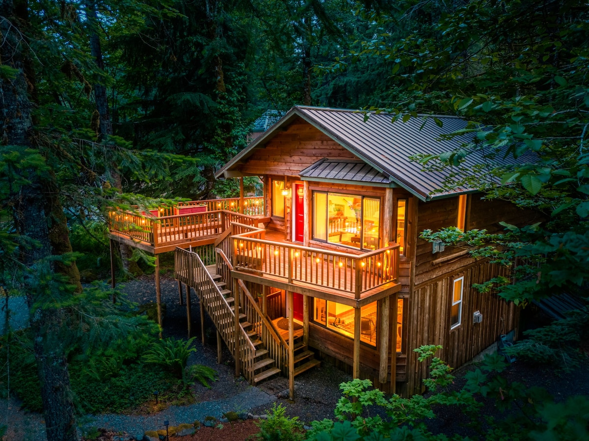 The cabin is nestled among trees, featuring a wooden facade with multiple decks. Warm light spills from large windows, highlighting the inviting interior. A staircase leads to the upper deck, providing access to outdoor seating areas surrounded by lush greenery.