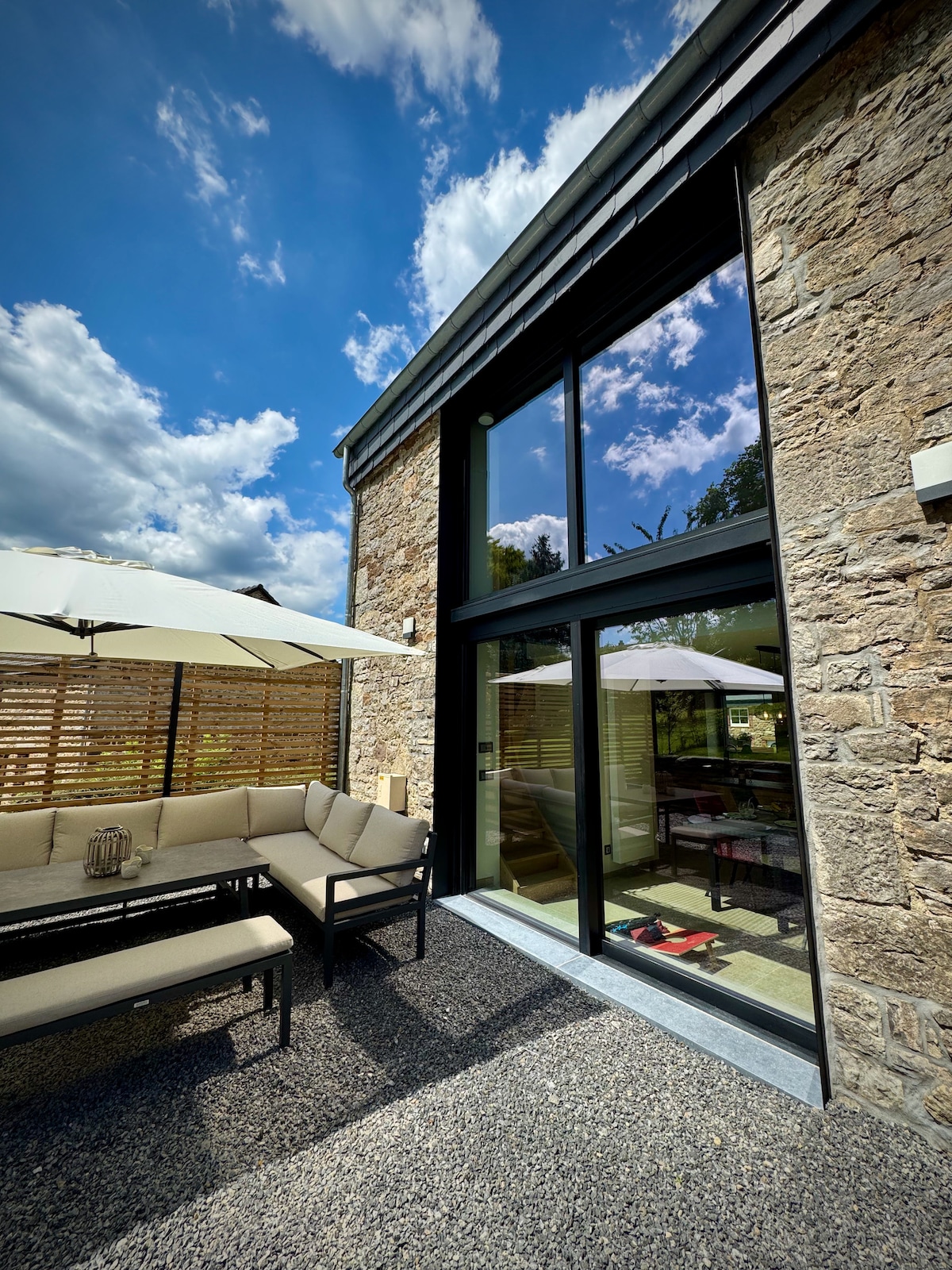 A spacious outdoor seating area is visible, featuring a comfortable sectional sofa positioned on a gravel surface. Large glass doors provide a view of the interior, reflecting surrounding greenery under a bright sky with scattered clouds. Umbrellas offer shade, enhancing the inviting atmosphere.