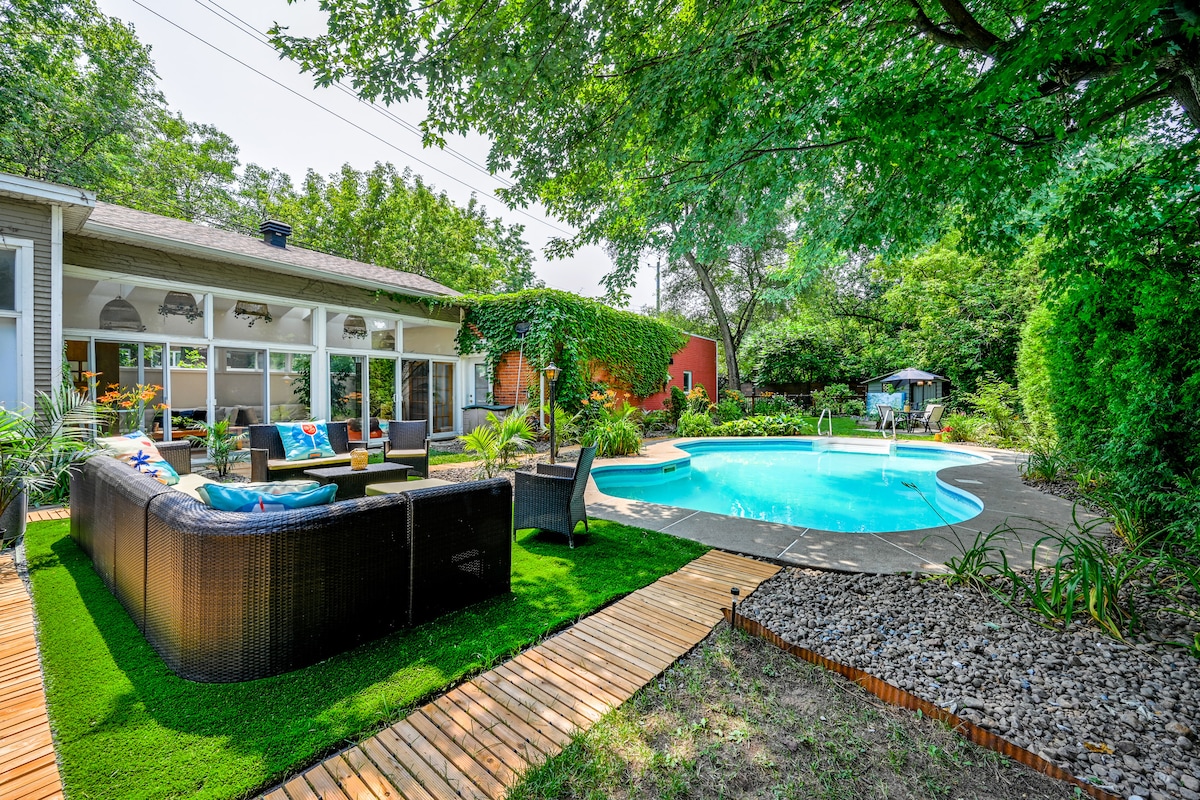 A serene backyard space showcases a heated outdoor pool surrounded by lush greenery. Comfortable seating in a cozy lounge area is set against a backdrop of vibrant landscaping. The scene invites relaxation and gathering, accentuated by natural light filtering through the surrounding trees.