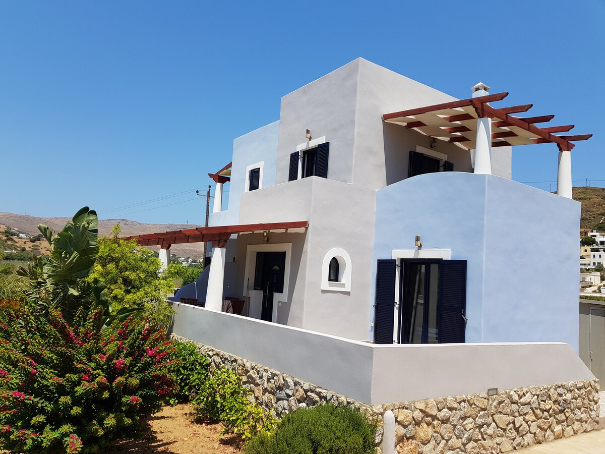 A two-story building is depicted, featuring a mix of light blue and white exteriors. The structure has several windows with dark shutters and a covered terrace, supported by wooden beams. Lush greenery surrounds the home, providing a contrasting burst of color.