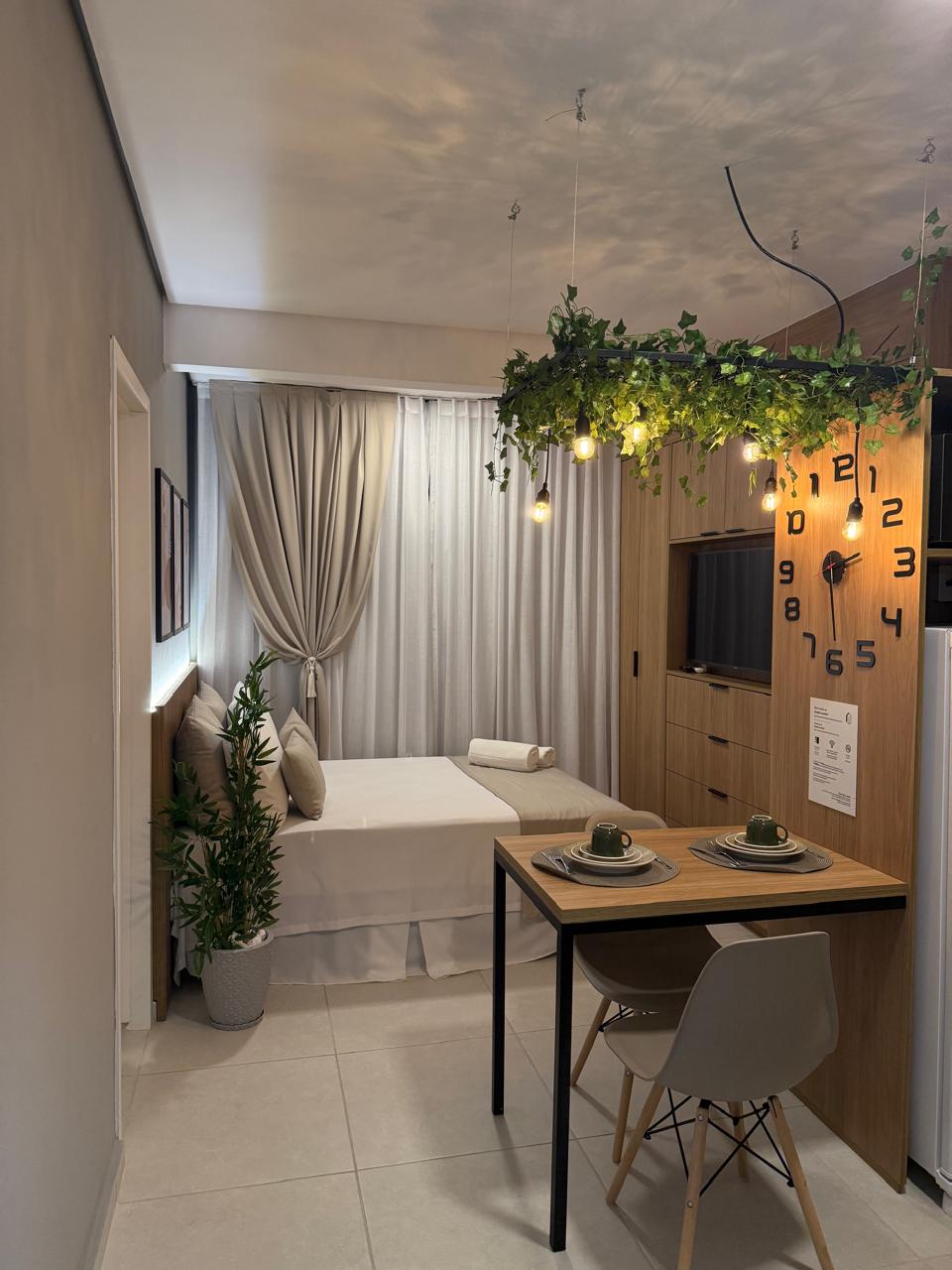 A compact studio space showcases a comfortable sleeping area fitted with a light-colored bedspread. A small dining table with two place settings occupies the center of the room. Decorative plants and soft lighting create a welcoming ambiance. Natural light filters through sheer curtains.