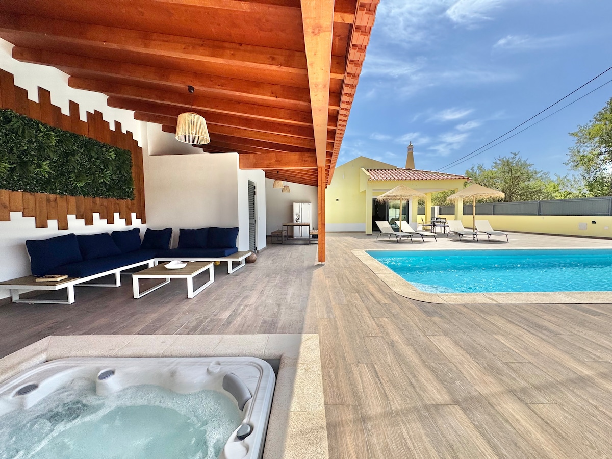A spacious outdoor area features a tranquil jacuzzi and a swimming pool surrounded by loungers. A shaded seating area with dark cushions offers relaxation. The wooden ceiling provides shelter, while clear blue skies add to the inviting ambiance.