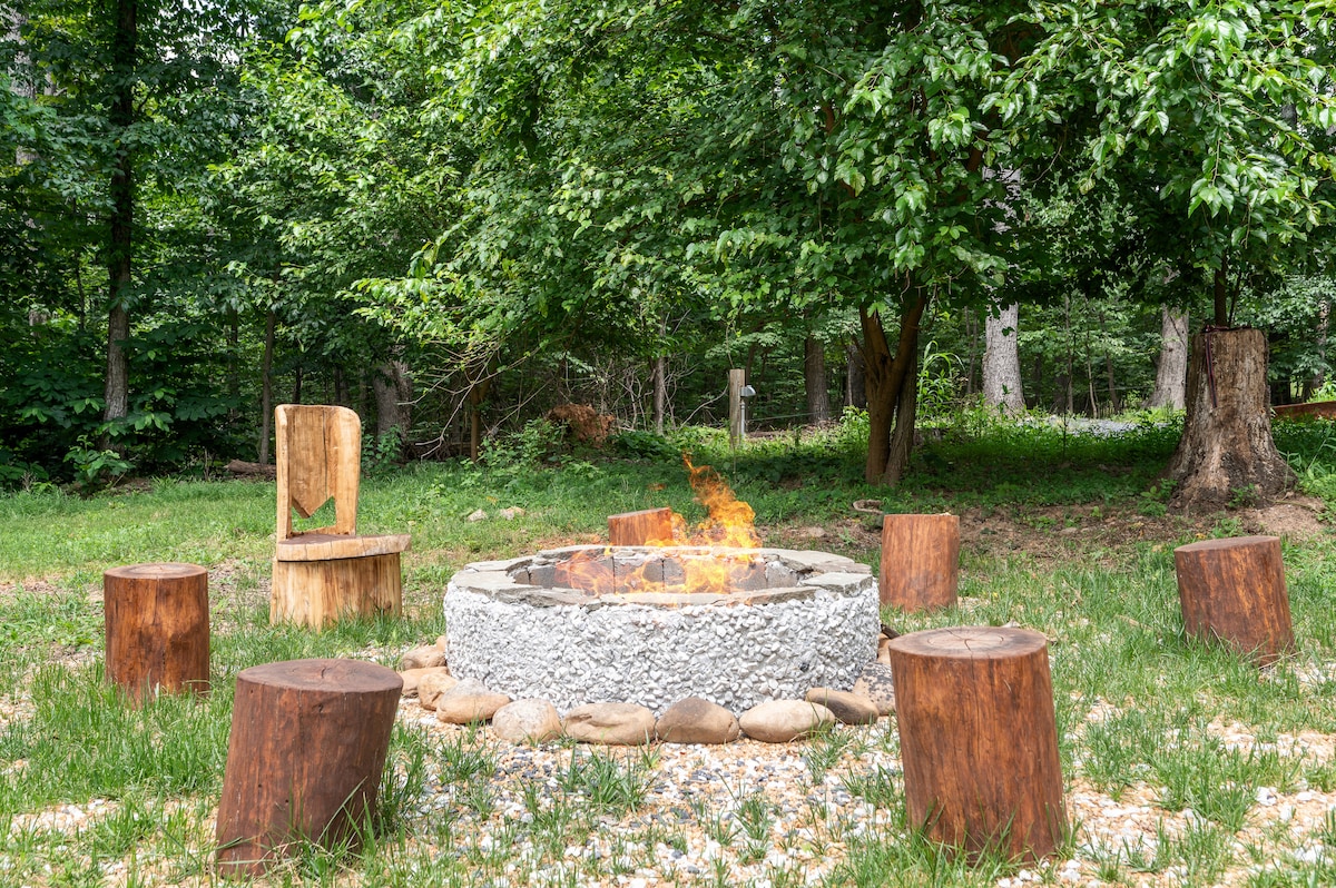 An outdoor fire pit is surrounded by natural stone seating, crafted from logs. Lush green grass covers the area, while trees provide a serene backdrop. A gentle flame burns in the center, inviting gatherings under the open sky.