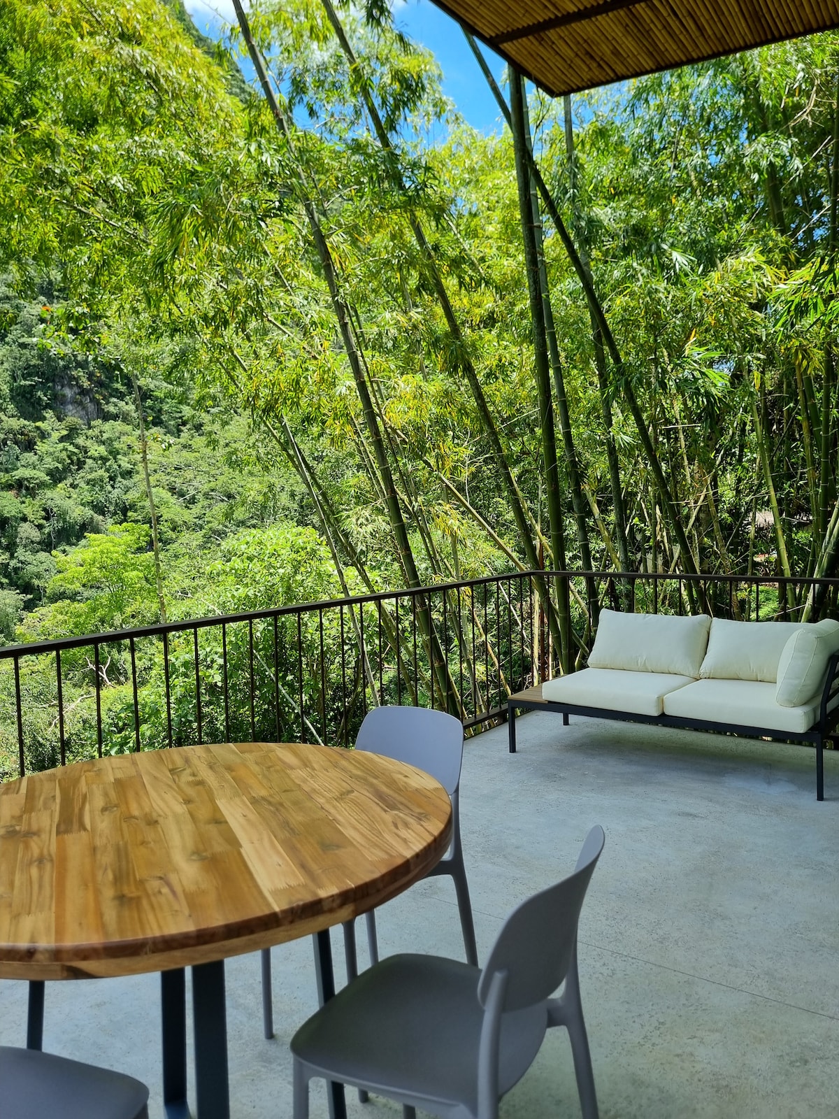 An outdoor terrace features a round wooden table surrounded by four light-colored chairs. A plush white sofa is positioned against the railing, providing a view of lush green bamboo and distant hills. The area is bathed in natural light, enhancing the serene atmosphere.