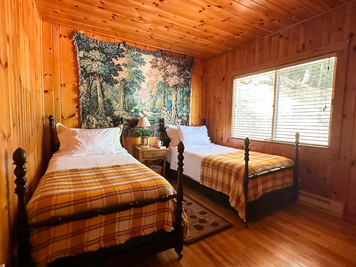 Tapestry Room with knotty pine walls, two antique twin beds, and a woodland wall hanging. A wide window offers views of the lake and gentle light. A bedside alarm clock with 2 USB ports (not shown in photo) has been added for convenience.