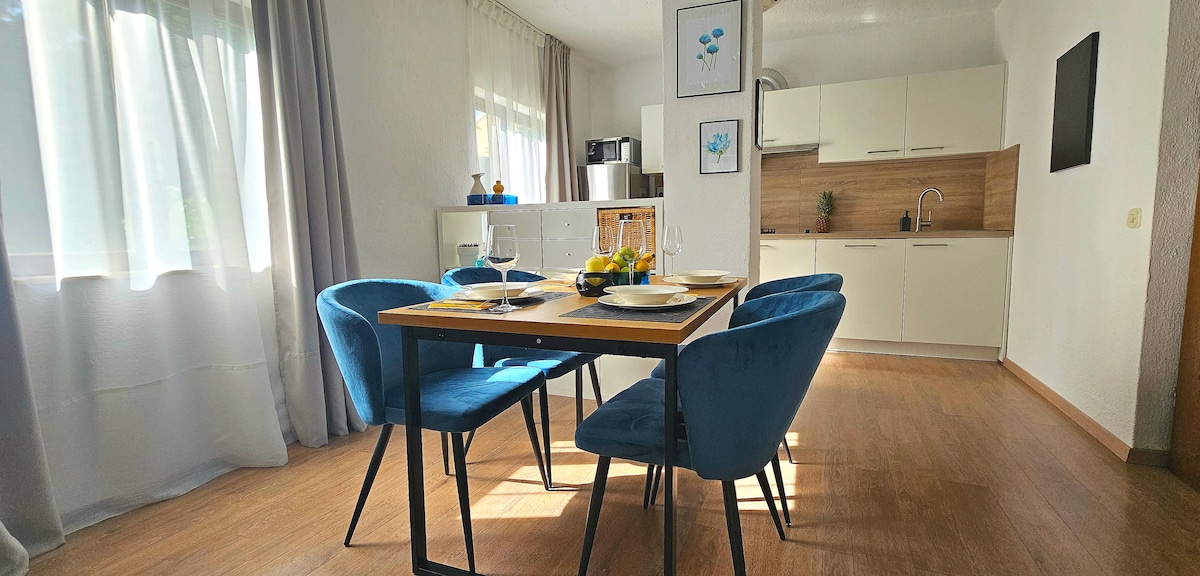 A modern dining area features a wooden table set for four, accompanied by plush blue chairs. Natural light filters through sheer curtains, brightening the space. A compact kitchen is visible in the background, showcasing sleek cabinetry and essential appliances.