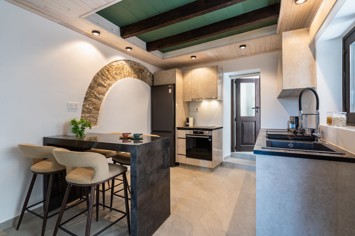 A modern kitchen blends stone and sleek finishes, featuring a dark countertop with four tall stools. Appliances include a refrigerator and an oven, with a window providing natural light. The ceiling showcases exposed beams, complementing the contemporary design.