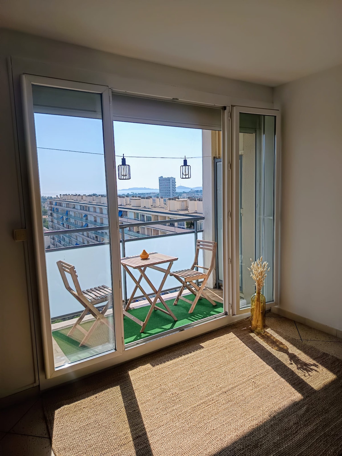 A balcony space is visible, featuring a small outdoor table with two wooden chairs. A clear view extends beyond the glass doors, showcasing the city with distant views of the sea. Soft light filters through, illuminating the interior decor.
