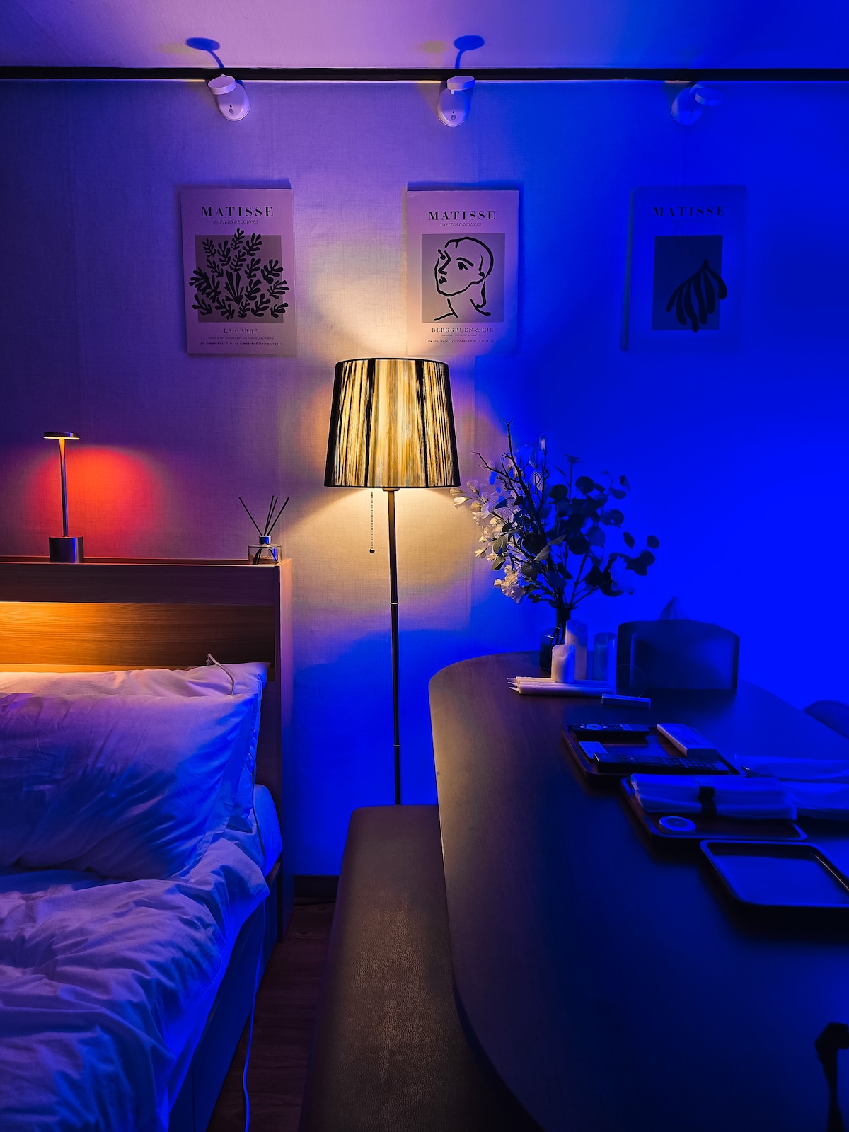 A cozy sleeping area features a queen-size bed with white bedding, complemented by warm lighting from a floor lamp. A modern table is visible, adorned with various items. Artistic posters are displayed on the wall, and colored lighting adds a calming ambience.