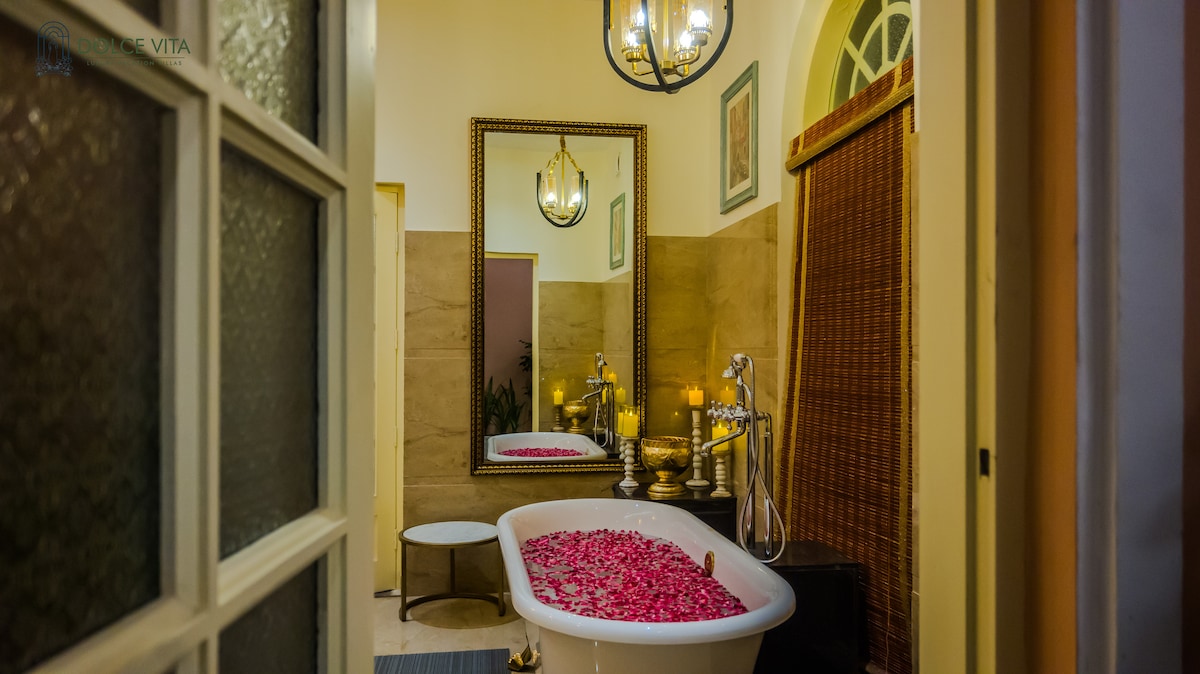 A luxurious bathroom showcases a freestanding bathtub filled with pink rose petals. A large mirror reflects the elegant fixtures, while warm lighting is provided by a chandelier. Natural elements are complemented by a woven privacy screen and a small seating area.