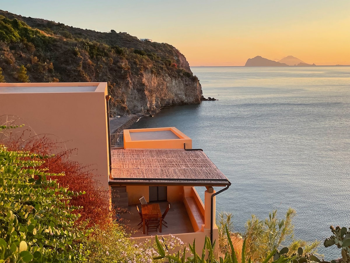The image captures a serene sunset over the sea, illuminating the exterior of a two-level house. A terrace is visible, furnished with wooden furniture, and lush greenery surrounds the property. In the background, distant islands can be seen across the calm waters.