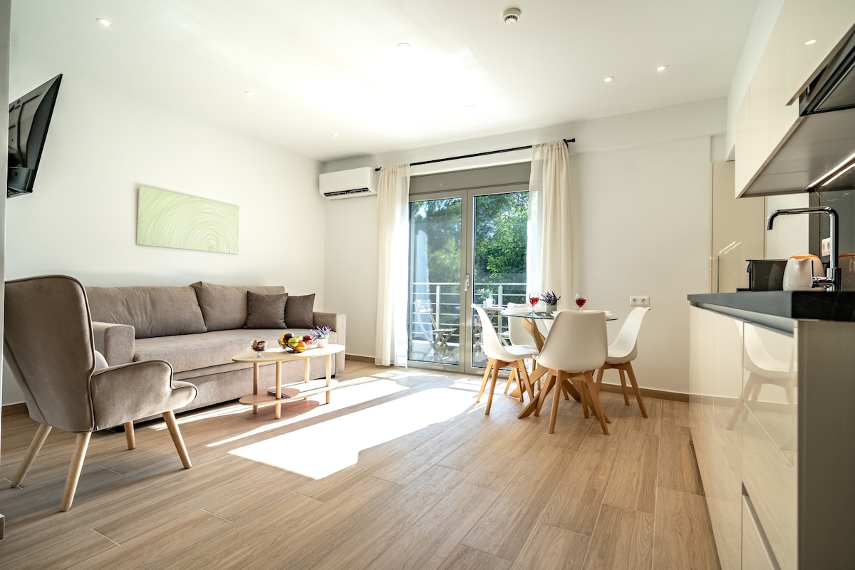 A modern living space is presented, featuring a light grey sofa and a coffee table with fresh fruit. A dining area with white chairs is visible near large glass doors that lead to a balcony. Natural light fills the room, highlighting its minimalistic decor.