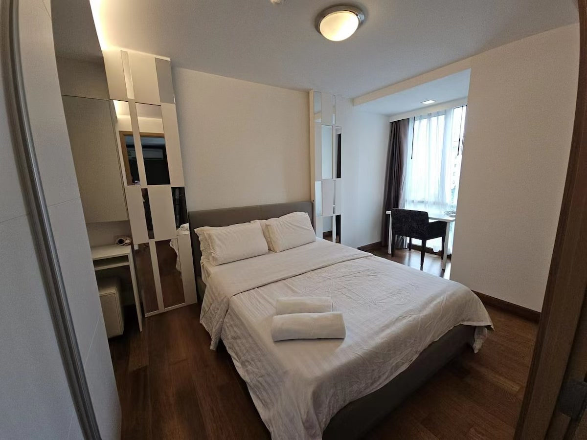 A spacious bedroom features a comfortable bed with multiple pillows and neatly arranged towels. A mirror accent wall reflects light, adding depth to the room. A window provides natural illumination, and a small desk with a chair is positioned nearby.