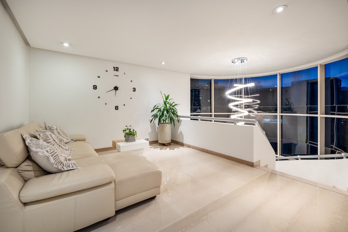 Este espectacular salón destaca por su doble altura y diseño moderno que combina amplitud, luminosidad y confort. Está amoblado con sofás blancos de gran tamaño, perfectos para compartir en grupo o relajarse viendo televisión.