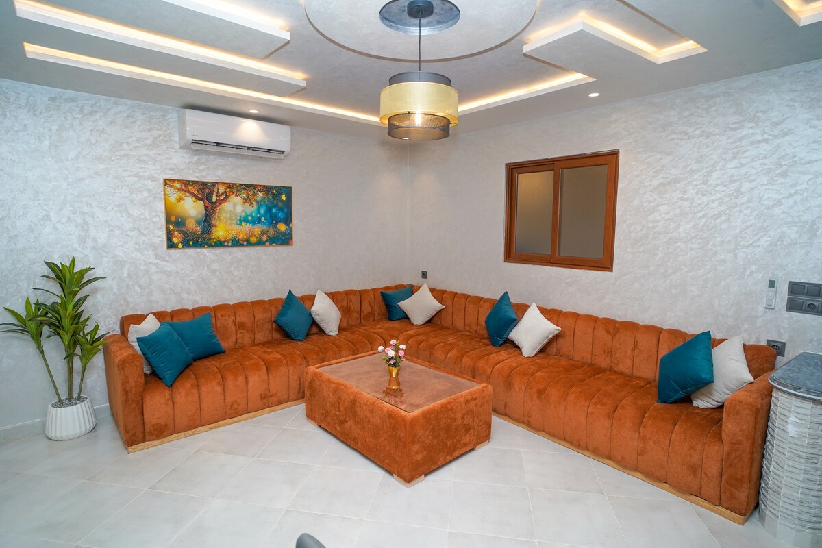 A spacious living area features an orange sectional sofa adorned with teal accent pillows. A central coffee table is positioned atop white tiles, with a small flower arrangement placed in the center. Ambient lighting is provided by a modern overhead fixture, and a window allows natural light to fill the space.