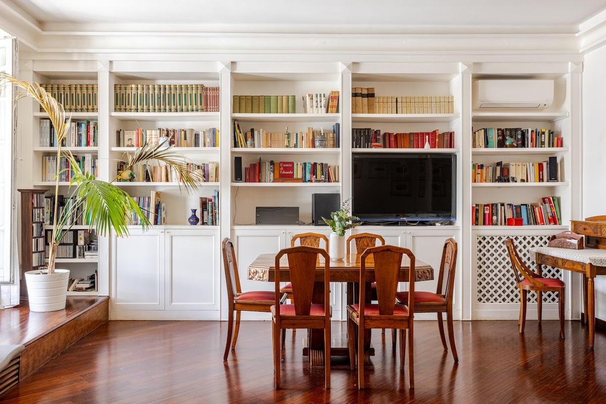 A spacious living area features extensive bookshelves filled with various books, complemented by a large television. A dining table is surrounded by four elegant chairs, and a decorative plant adds a touch of greenery to the space. Warm wood flooring enhances the inviting ambiance.