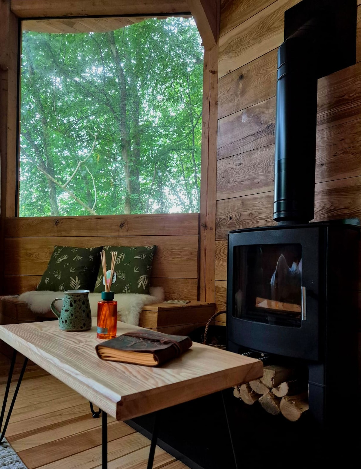 A cozy corner of the cabin features a modern wood stove surrounded by natural wood paneling. A wooden coffee table stands in front of a comfortable seating area adorned with leafy patterned cushions. Large windows invite views of the lush greenery outside.