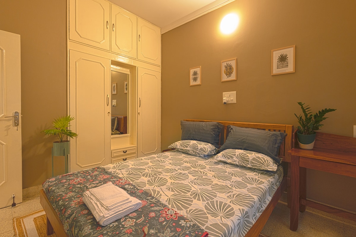 A cozy double bedroom features a wooden bed covered with a patterned duvet and arranged pillows. The room includes a large wardrobe, bedside lamp, and a small workspace with a plant. Light is provided by a wall-mounted fixture, creating a calming atmosphere.
