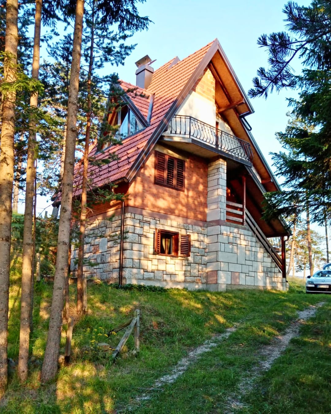 A charming mountain cabin is framed by tall pine trees. The structure features a combination of natural stone and wood with a sloped roof. A balcony is visible on the upper level, overlooking the surrounding greenery.
