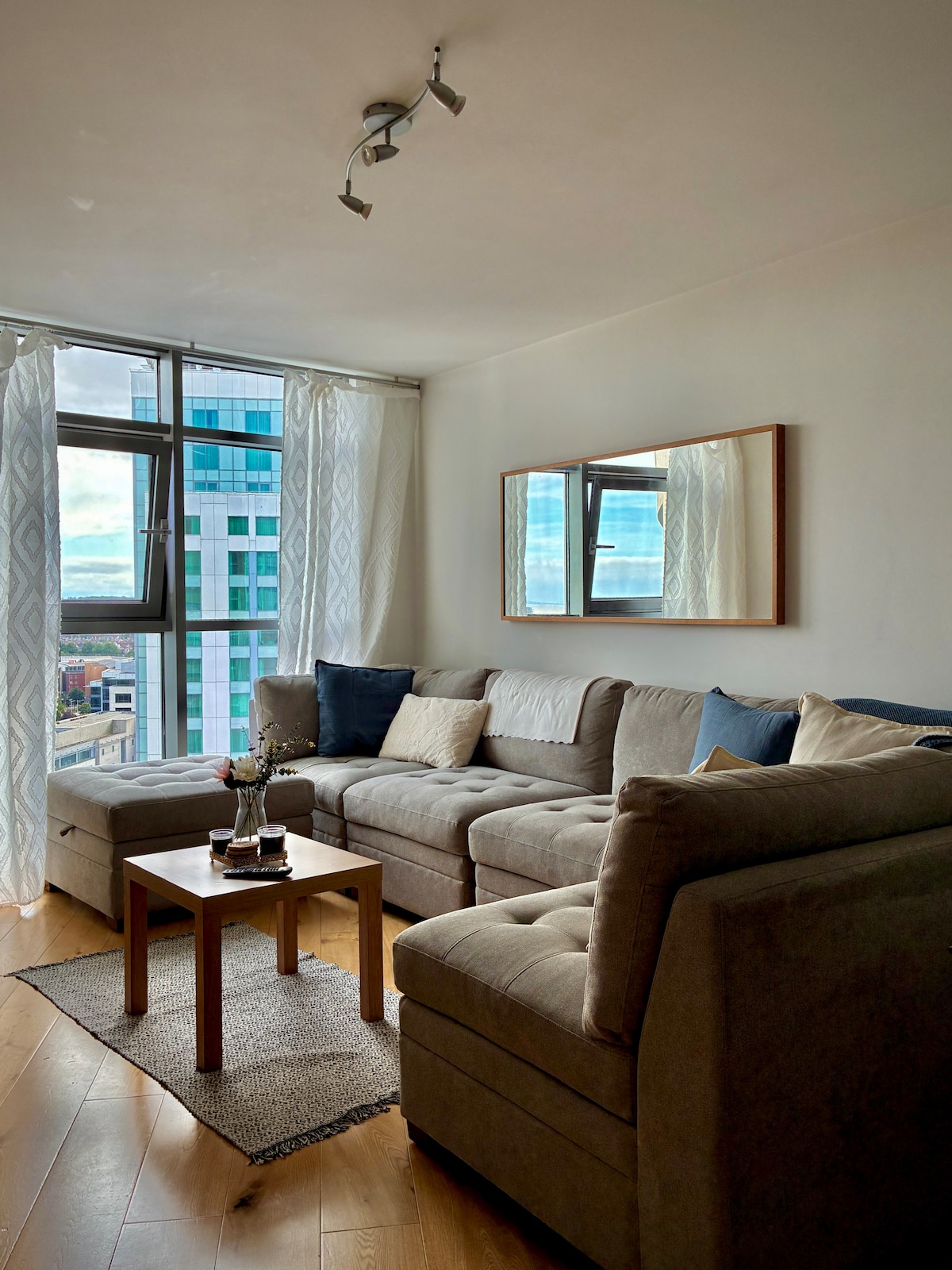 A spacious living area is presented with a large sectional sofa adorned with blue and cream pillows. A small wooden table sits in front of the sofa, positioned on a woven rug. Two tall windows with light curtains allow natural light to fill the space.