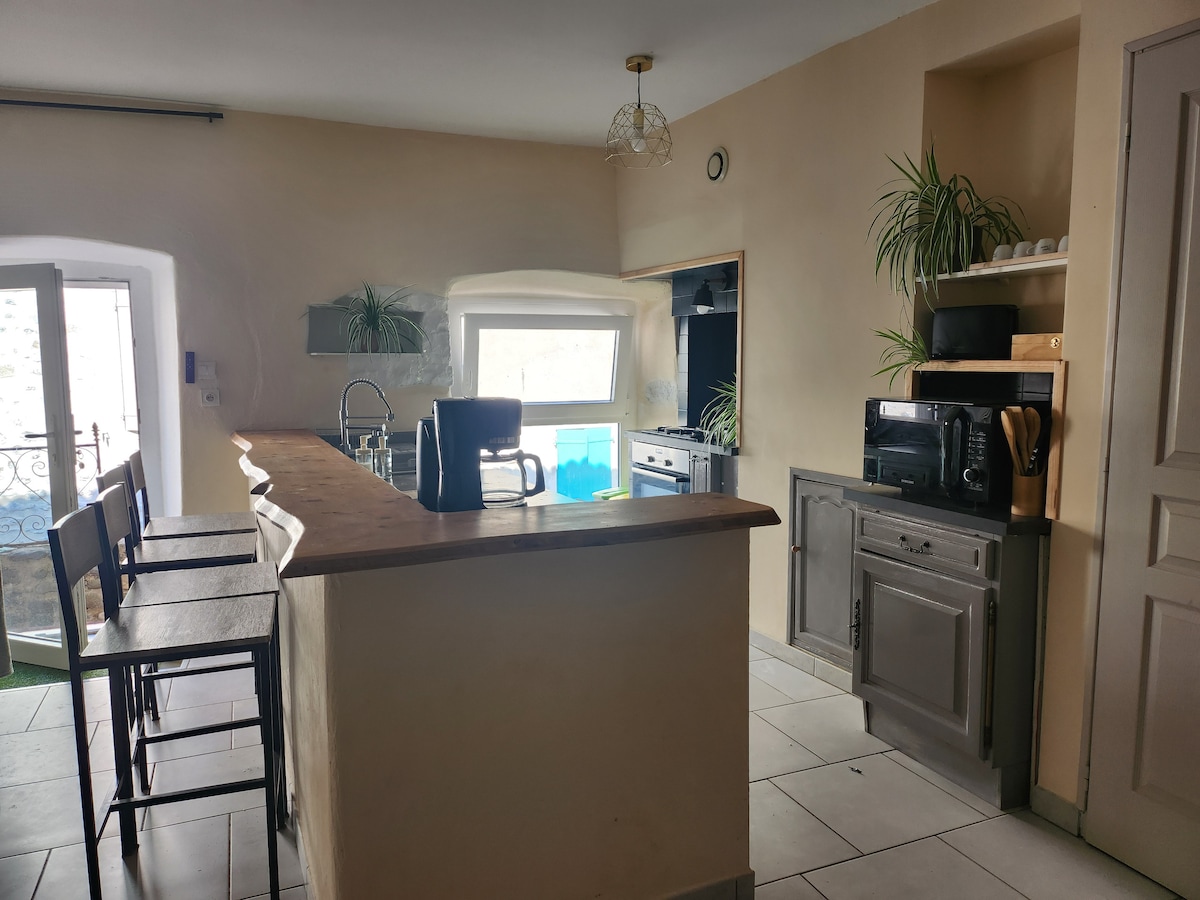 A modern kitchen features a wooden countertop along with high-backed stools. Plants are displayed on shelves, and appliances include a microwave, oven, and sink. A window above the sink offers a glimpse of the outdoor area, enhancing natural light within the space.