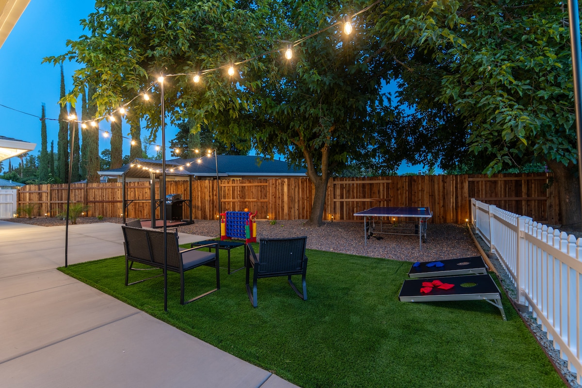 A spacious backyard is depicted, featuring a well-manicured lawn and string lights illuminating the area. Comfortable seating is provided around a small fire pit, with a colorful game set nearby. A ping pong table is also visible, emphasizing recreational opportunities.