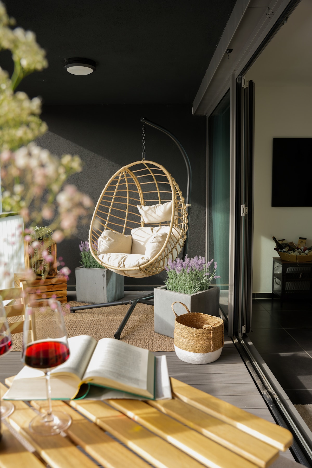 A cozy balcony area features a hanging chair with soft cushions and a decorative potted plant. A wooden slatted table holds a glass of red wine and an open book. Sunlight gently illuminates the space, inviting relaxation and comfort.