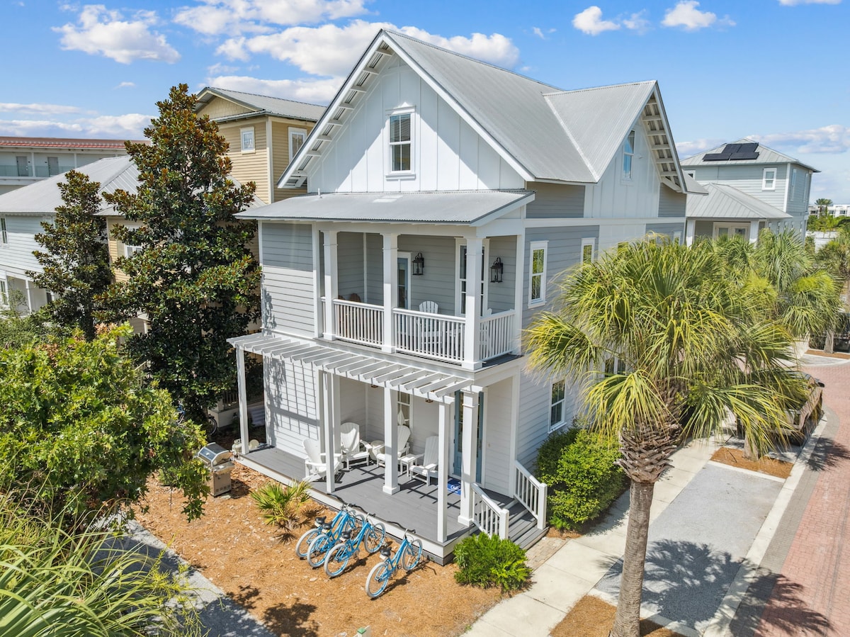 A charming three-story beach home is showcased with a spacious front porch. The entrance is framed by vibrant foliage and palm trees. Four bicycles are neatly lined up on the driveway, providing options for exploration. The exterior features light blue siding under a metal roof.