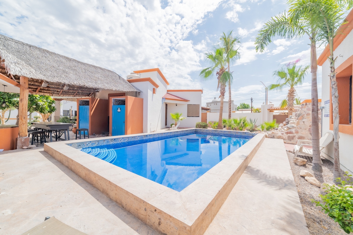 A serene pool area is highlighted by a gleaming blue swimming pool, surrounded by tropical palm trees and desert landscaping. A shaded palapa with seating and a dining area offers a relaxing spot for outdoor gatherings, creating a calm and inviting atmosphere.