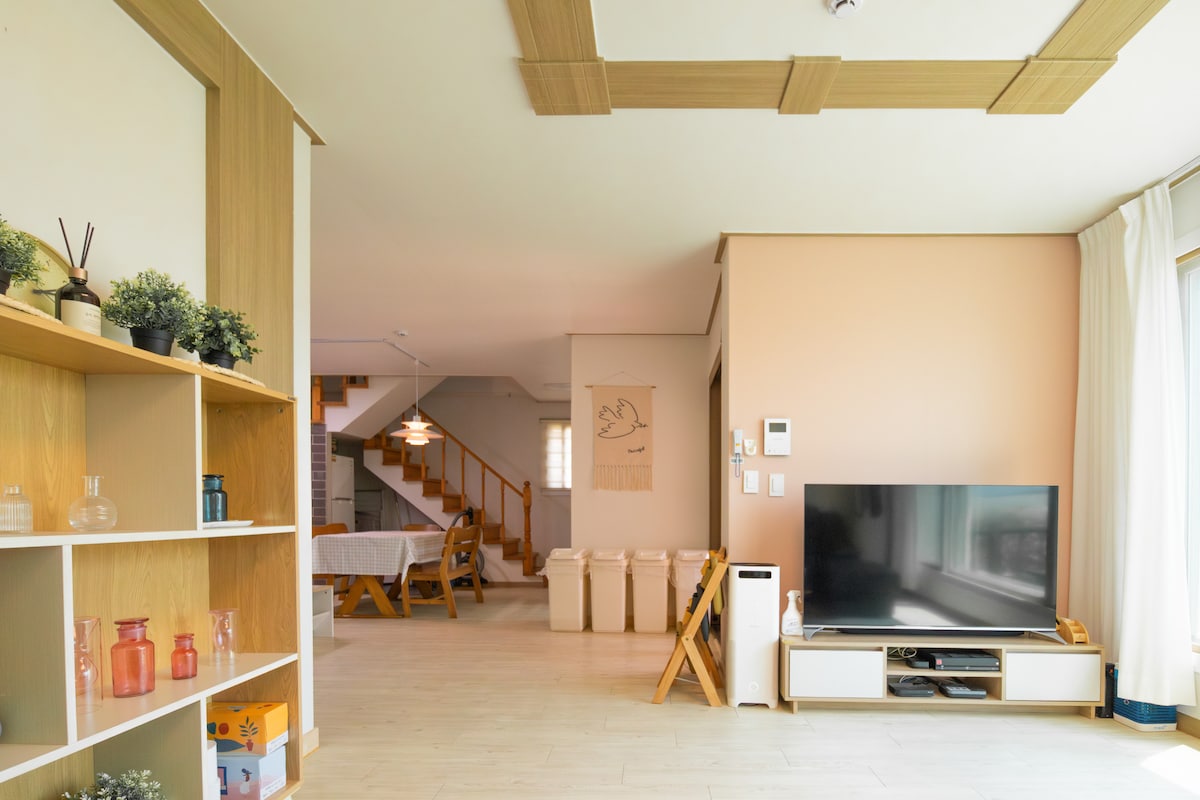 A spacious living area displays a flat-screen television on a low media console. Light-coloured walls and wooden accents create a calm environment. To the left, stairs lead to the upper level, while baskets and a cozy sleeping area are visible in the background.