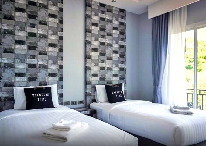 A bright room features two spacious beds covered with white linens, each accompanied by a folded towel. The walls are adorned with a modern, patterned design, and large windows allow natural light to brighten the space, revealing views of greenery outside.